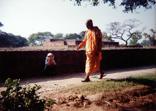 A monk dressed in orange robes walks by a young American boy visiting the location of Buddha's meditation garden at Shravasti, where Buddha spent many rainy seasons, bricks mark the old layout of the space, India by Wonderlane
