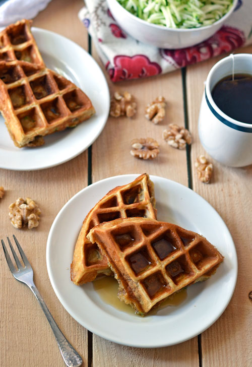 Two white plates with zucchini waffles and syrup next to a mug of tea and walnuts