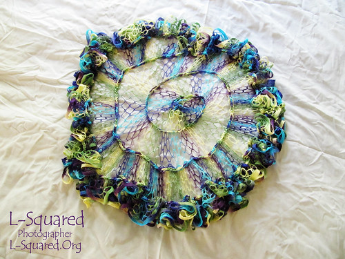 Circular, spiraled mesh shawl in hues of yellow, green, teal, blue and purple.