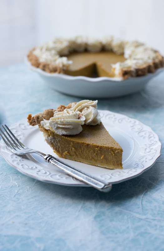 slice of pumpkin pie on small white plate with fork pumpkin pie in white pie dish in background with slice removed