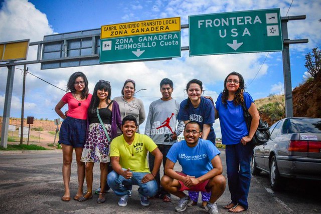 Eight immigrant youths stand in front of a highway sign that reads "frontera"