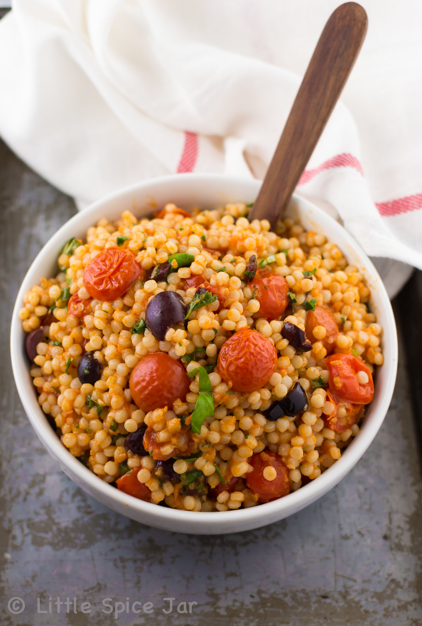 bowl of prepared Mediterranean pearl couscous salad on sheet pan with wooden spoon