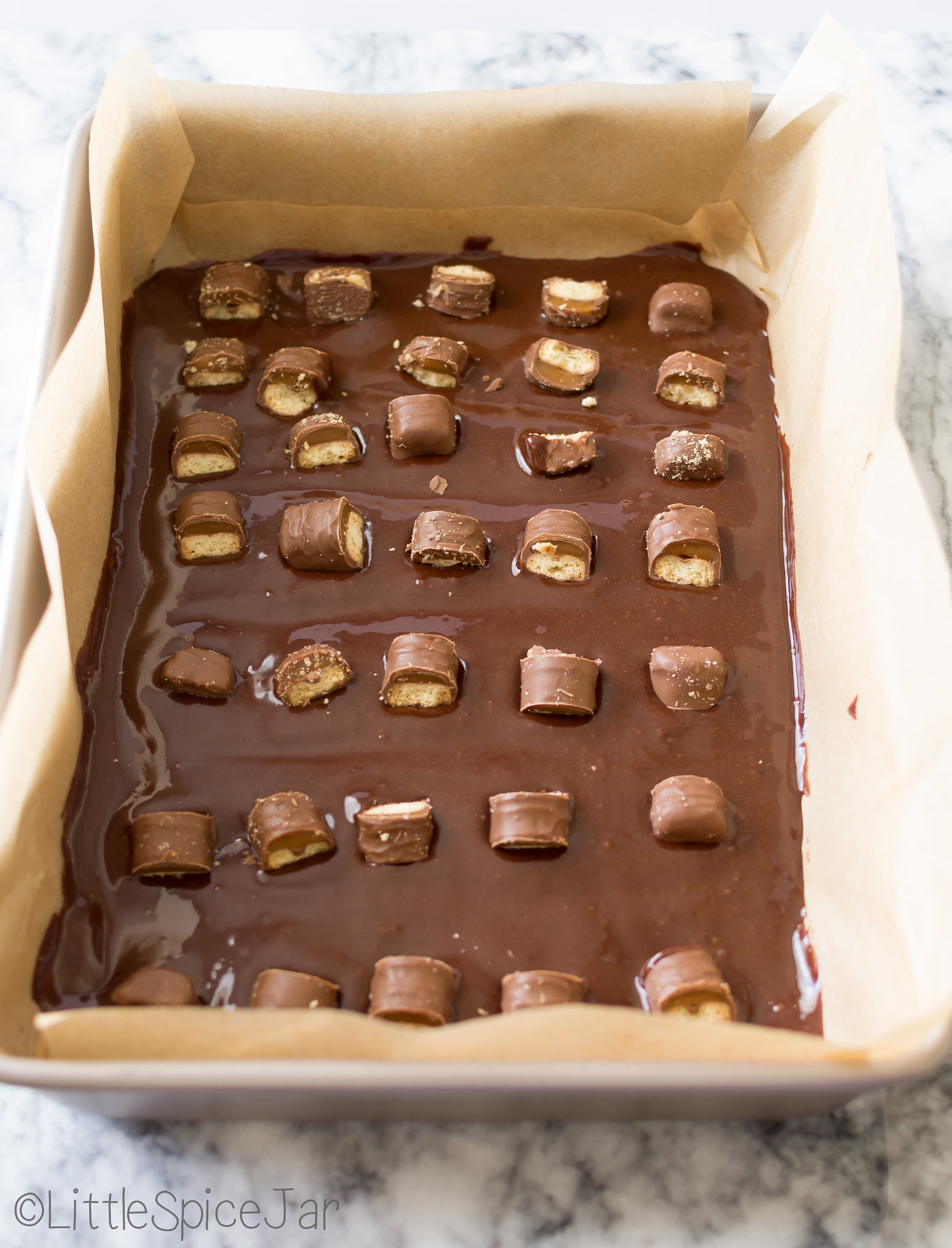 brownie batter topped with broken Twix candy bars on parchment lined baking dish
