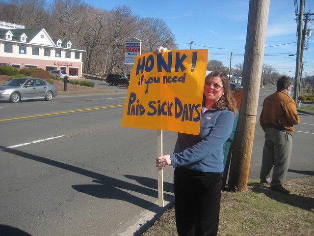 At Paid Sick Day Rally, Woman holds sign saying "Honk! if you need Paid Sick Days"