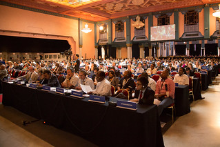 A large room with rows of table and chairs filled with diplomats from all over the world. On top of the tables are placards that display the country or organization that the diplomat is representing.