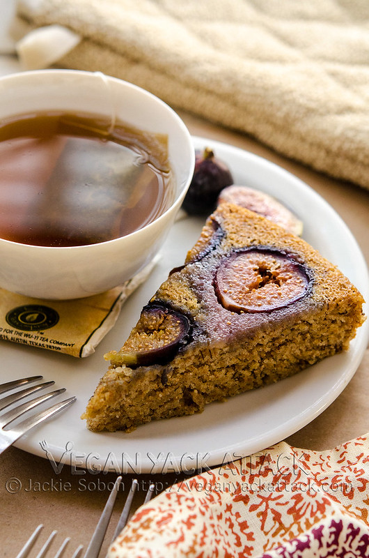 A slice of vanilla cake with figs baked into it on a plate with a cup of tea and colorful linen