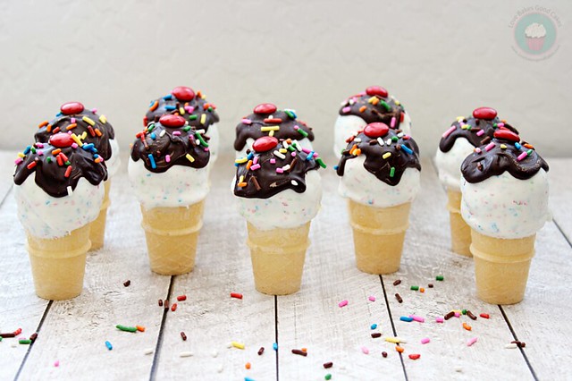 Ice Cream Cone Cake Pops with chocolate syrup and rainbow sprinkles standing up on counter with rainbow sprinkles.