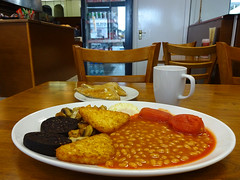 A cooked breakfast served on a large white plate.  Half the plate is taken up by a lake of baked beans.  A slice of toast, cut in half and heavily buttered, is on a smaller plate beyond, and a white mug sits to one side.  The table top is wood-effect formica.  A closed white-painted door and a fridge of soft drinks are beyond the table to the right, while to the left is a wooden service counter.  A wire basket holding various-coloured packets of crisps is on top of the counter.
