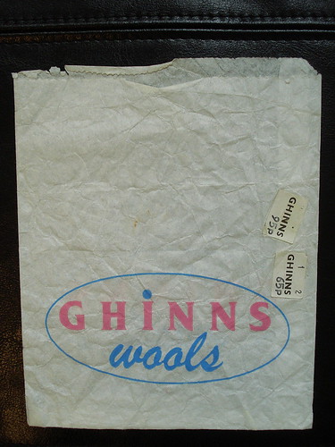 A paper bag with “Ghinns Wools” printed near the bottom. “Ghinns” is in light pink capital letters with a light blue dot over the “i”, and “wools” is in the same light blue in a cursive lowercase font.  Two small price tags are stuck to the bag, one reading “GHINNS 95p” and the other reading “GHINNS 65p”.