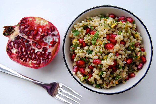 Israeli Couscous with Pomegranate & Pistachio in a white bowl with a black rim on a white background with a halved pomegranate.